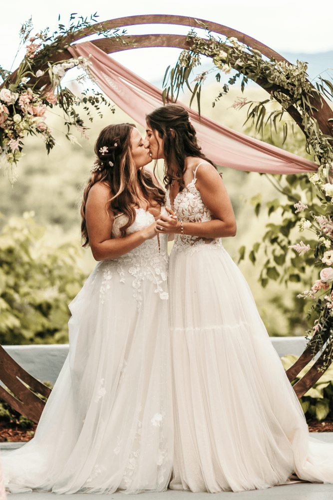 Two brides share a romantic kiss in front of a floral arch at their wedding ceremony.