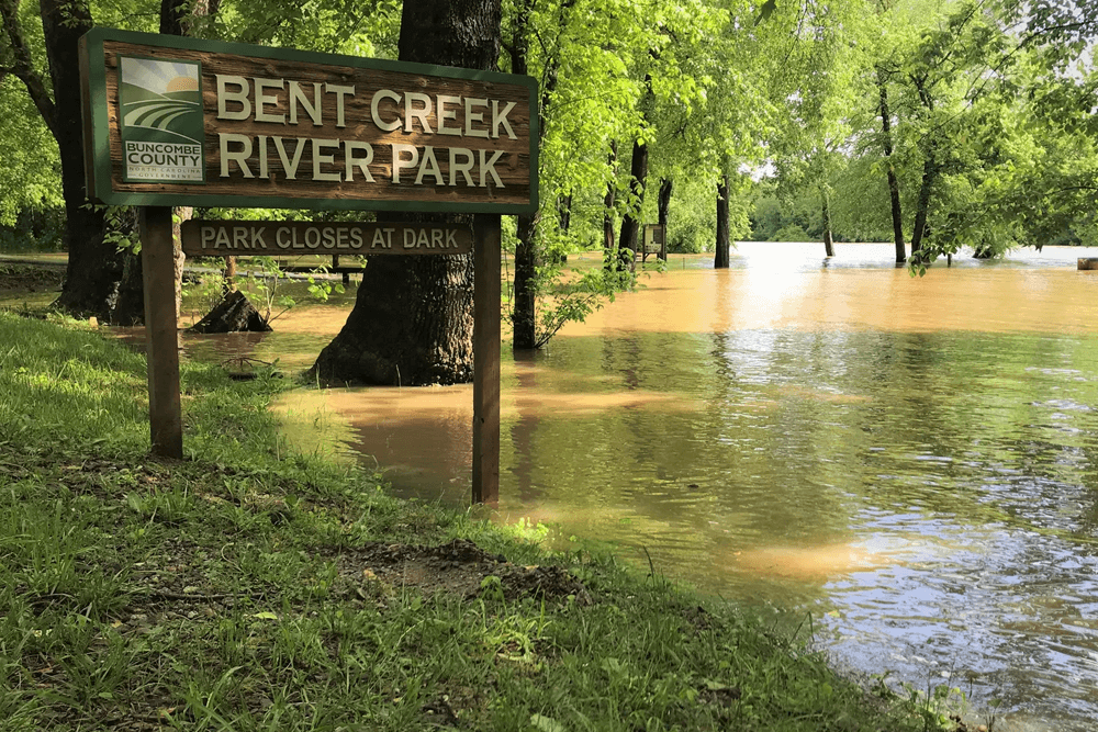 A wooden sign for Bent Creek River Park is partially submerged in floodwaters surrounded by trees.