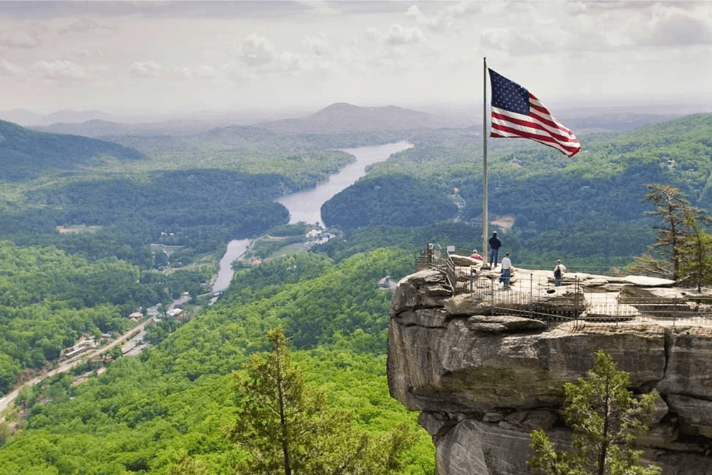 A panoramic view from a cliff with an American flag, overlooking a lush green valley and winding river.