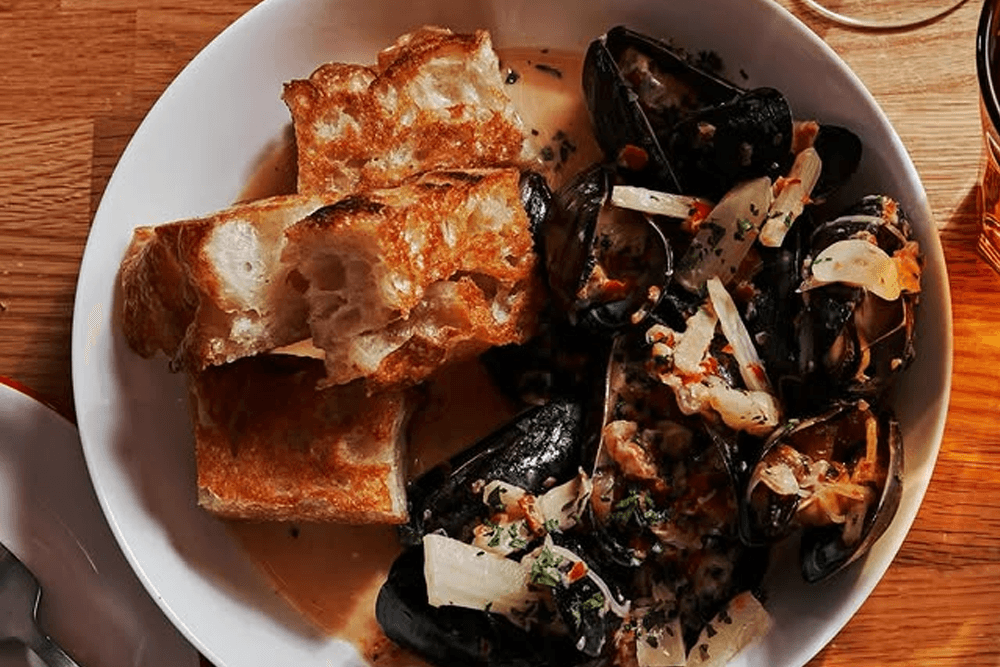 A bowl of mussels in broth with sautéed onions and herbs, accompanied by toasted bread.