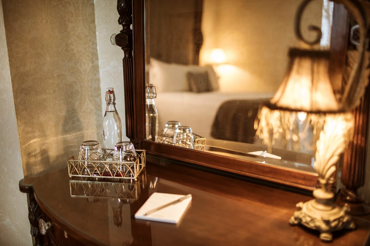 A wooden dresser with a mirror, featuring a lamp, water bottles, glasses, and a notepad.