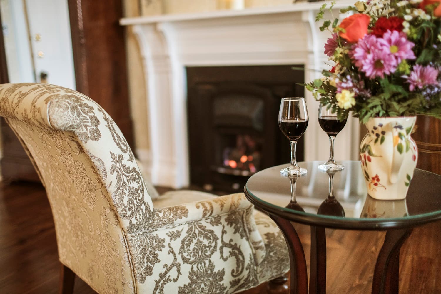 A cozy living room scene featuring a patterned armchair, a table with two glasses of wine, and a vibrant flower arrangement near a glowing fireplace.