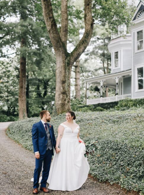 A bride and groom hold hands and smile at each other on a gravel path near a historic house surrounded by trees.