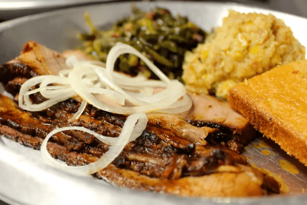 A plate with sliced brisket, topped with onions, served with collard greens, cornbread, and a scoop of grain dish.