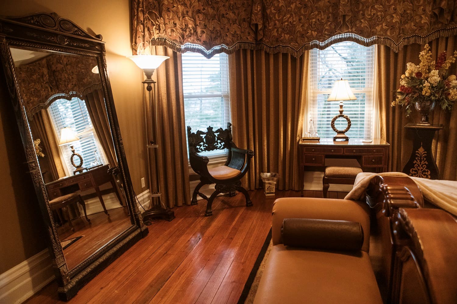 A cozy room featuring an ornate mirror, a wooden chair, a desk, and large windows with decorative curtains.