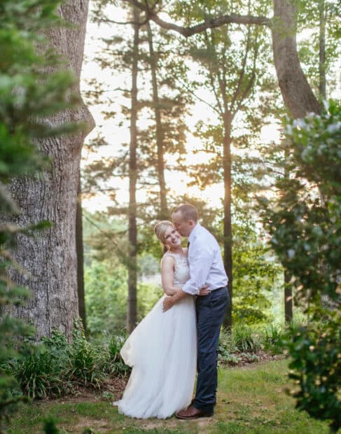 A couple embraces lovingly in a serene, wooded setting during sunset.