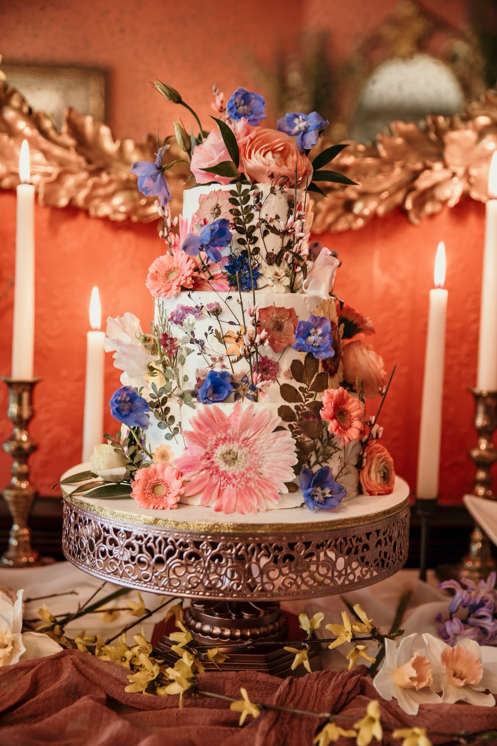 A beautifully decorated three-tier cake adorned with vibrant flowers sits on a decorative stand, surrounded by lit candles and soft fabric.