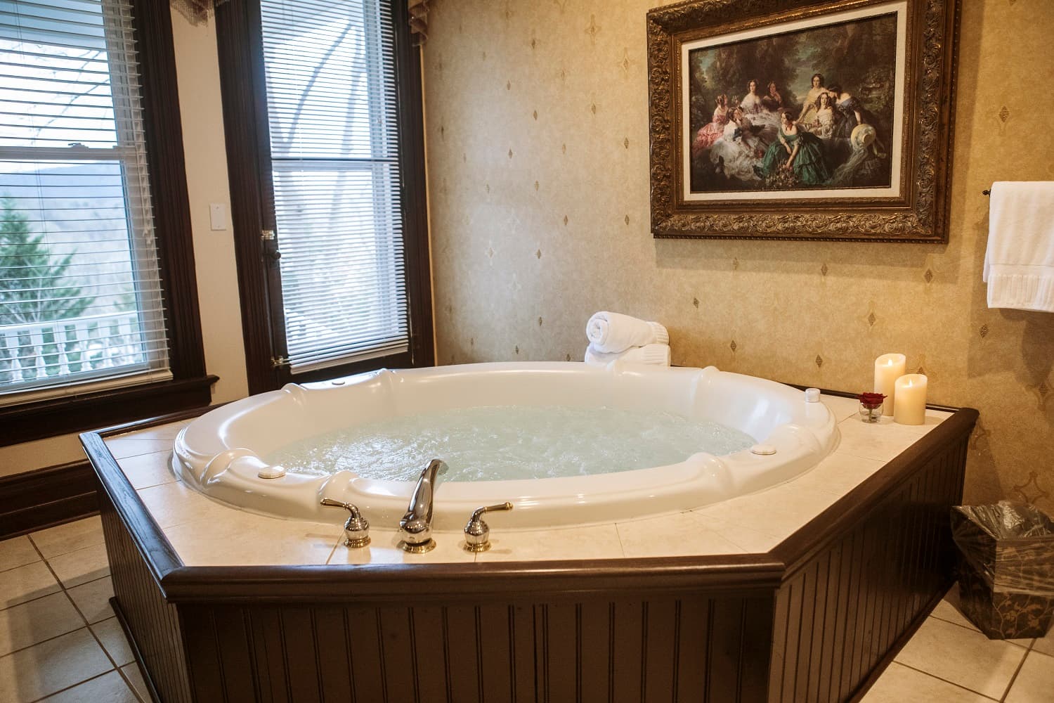 A white whirlpool bathtub is set in a warmly decorated bathroom with a large window and a framed painting on the wall.