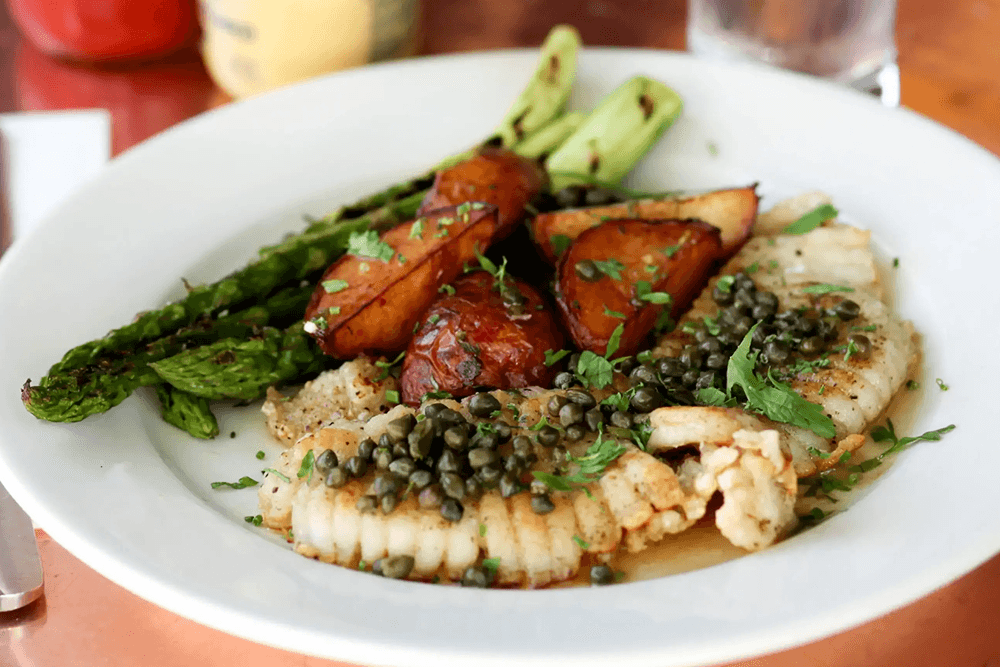 A plate of grilled fish with capers, alongside roasted potatoes and asparagus, garnished with fresh herbs.