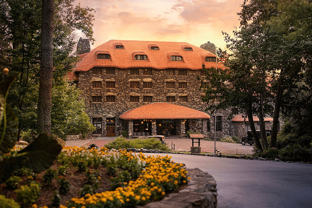 A rustic stone hotel with a red-tiled roof surrounded by lush greenery and flowers at sunset.