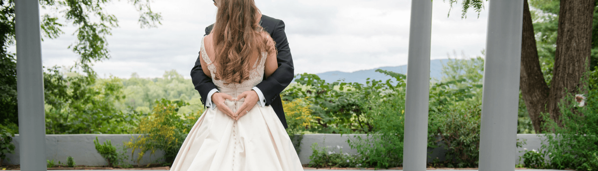 A couple in wedding attire embraces, forming a heart shape with their hands against a scenic backdrop.