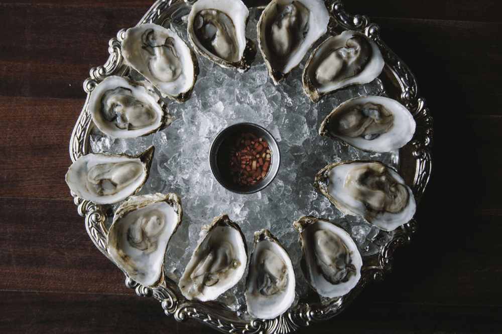 A platter of fresh oysters on ice, accompanied by a small bowl of sauce.