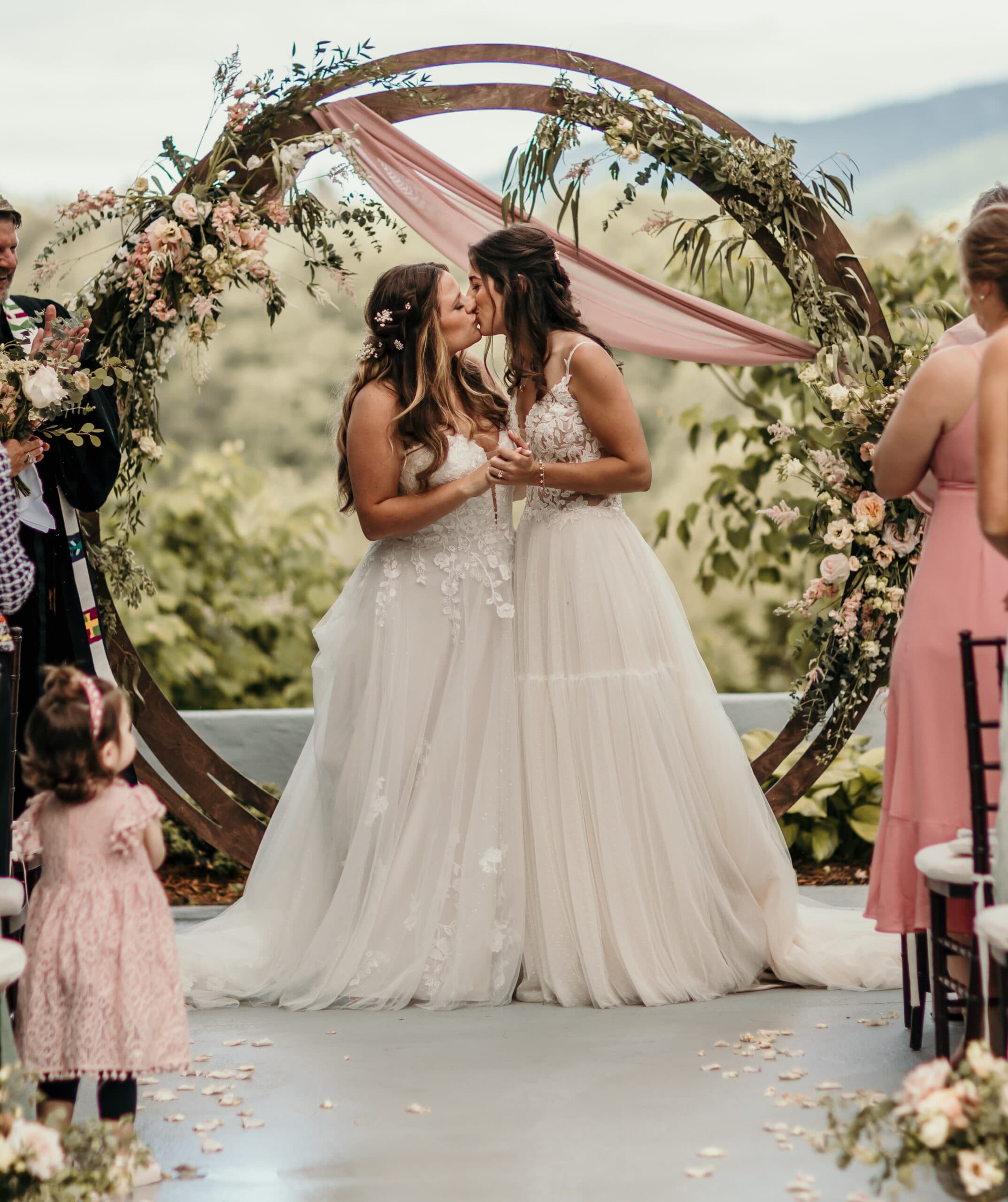 Two brides share a kiss under a floral arch during their wedding ceremony.