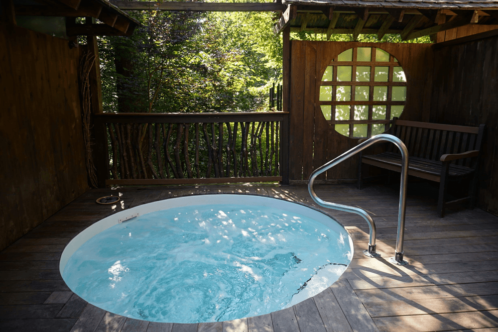 A wooden hot tub with bubbling water is situated on a terrace surrounded by trees.