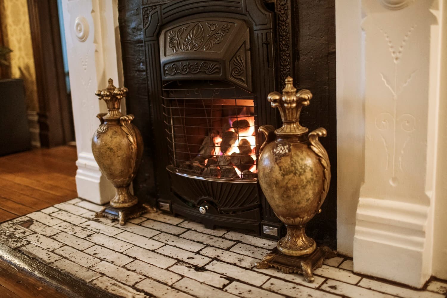 A decorative fireplace with a roaring fire flanked by ornate golden vases on a tiled hearth.