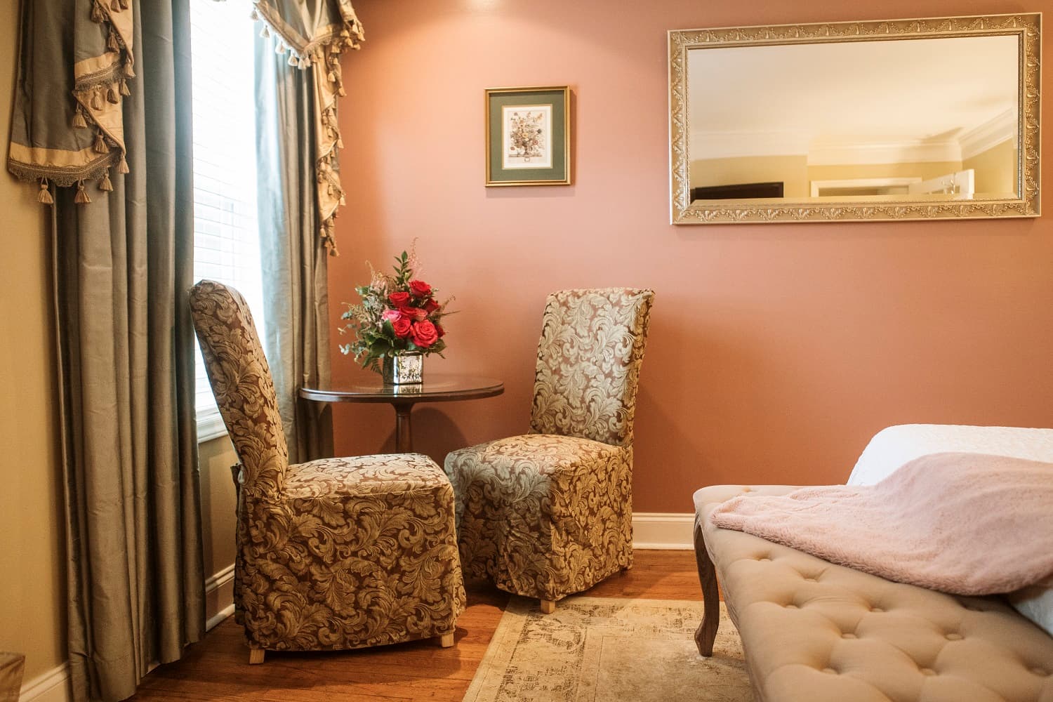 Cozy corner of a room featuring two patterned chairs, a small table with flowers, and a mirror on a warm-colored wall.