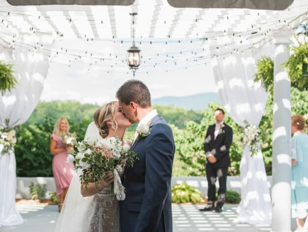 A bride and groom kiss under a decorative pergola during their wedding ceremony.