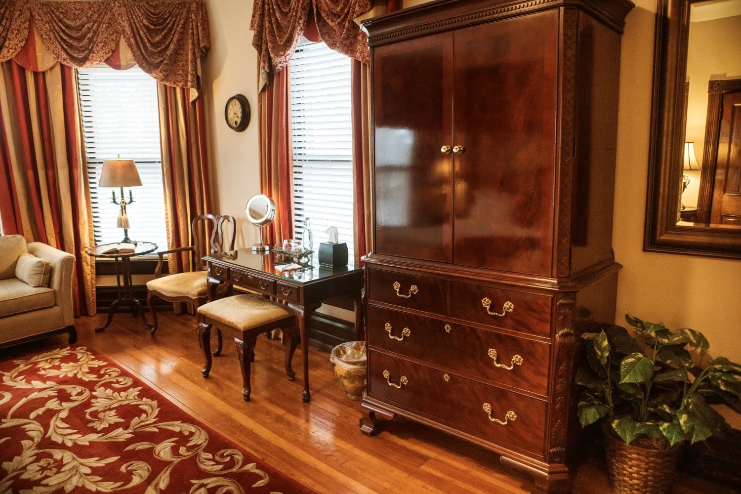 A cozy room featuring a wooden armoire, a small desk with a mirror, and a potted plant, complemented by elegant window treatments and a patterned rug.