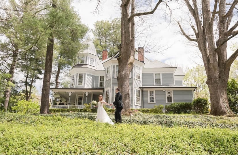 A bride and groom hold hands in front of a large, picturesque gray house surrounded by trees.