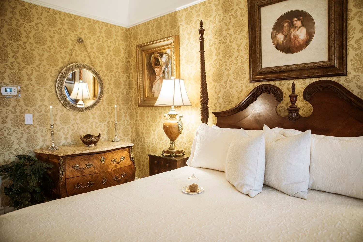 A cozy, elegantly decorated bedroom featuring a large bed with white pillows, antique furniture, and vintage artwork on the walls.