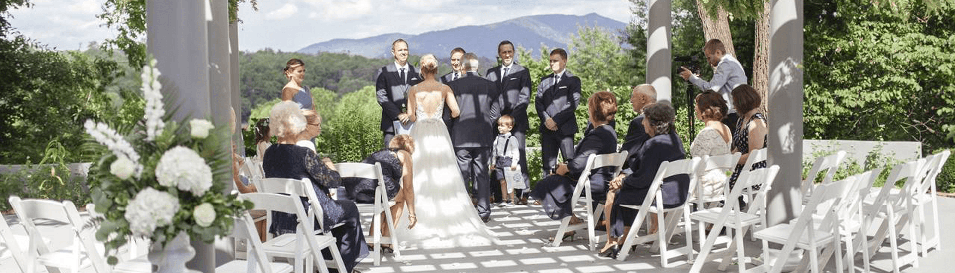 A bride and groom stand at the altar surrounded by wedding guests and a scenic mountain backdrop.