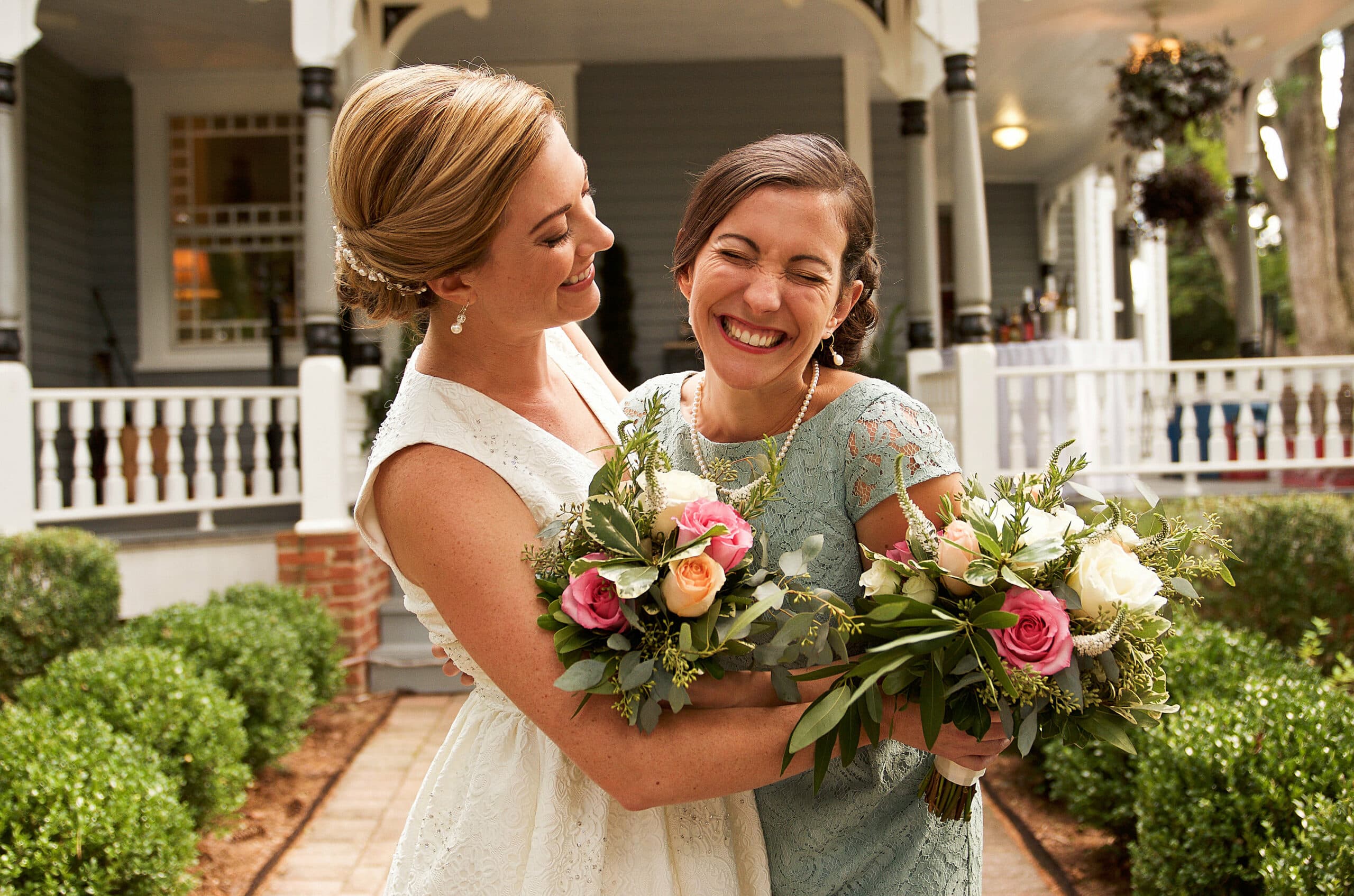 Two smiling women, one in a white dress and the other in a light blue dress, embrace while holding vibrant floral bouquets.