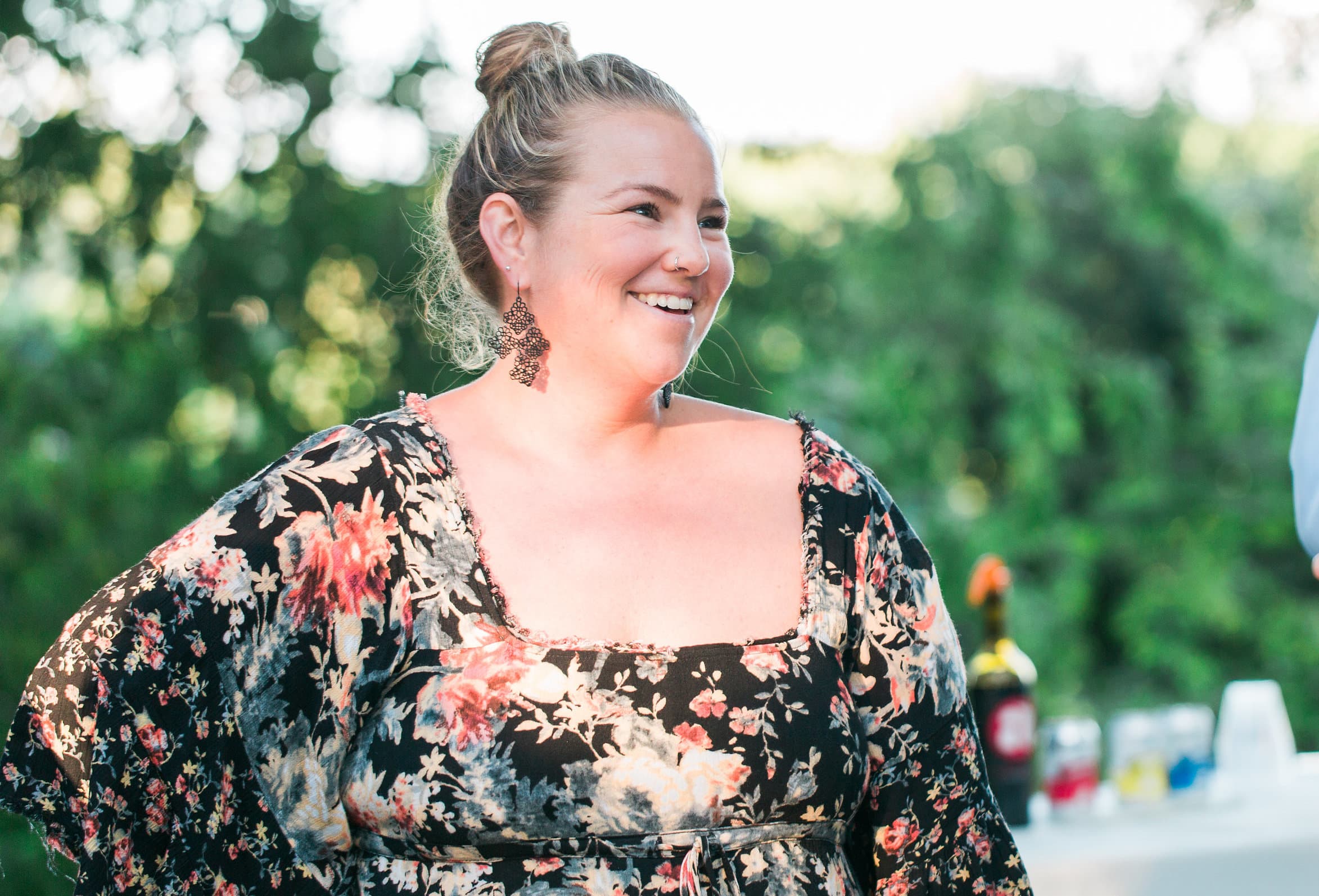A smiling woman in a floral dress stands outdoors with a blurred green background.