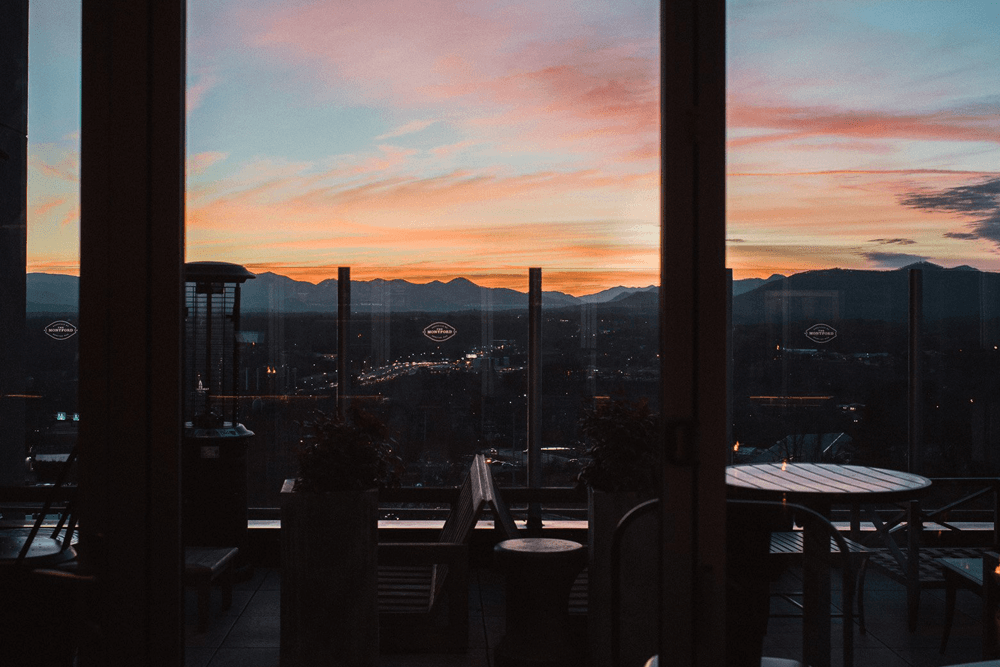 A sunset view over mountains is framed by glass doors and outdoor seating.
