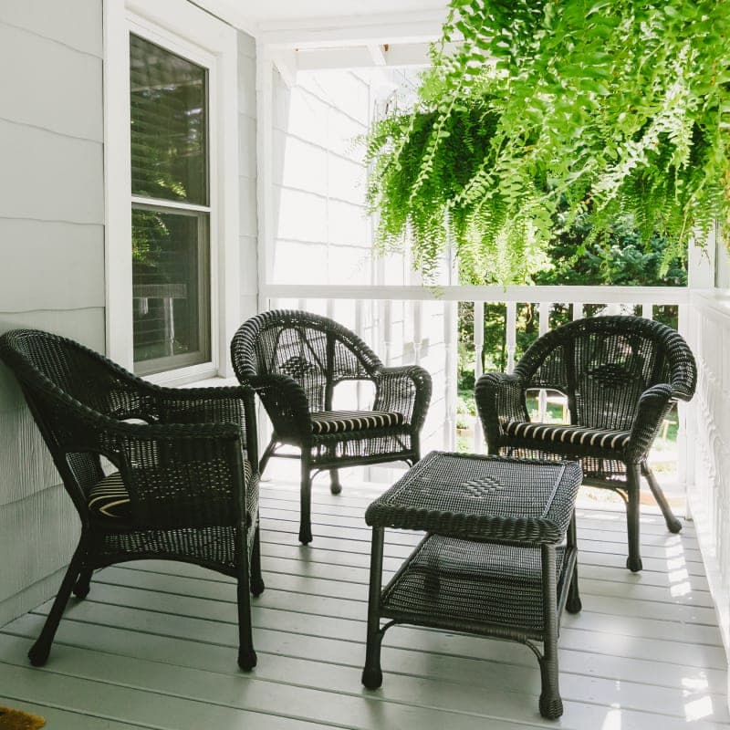 A cozy porch featuring four black wicker chairs and a small table, surrounded by green ferns.