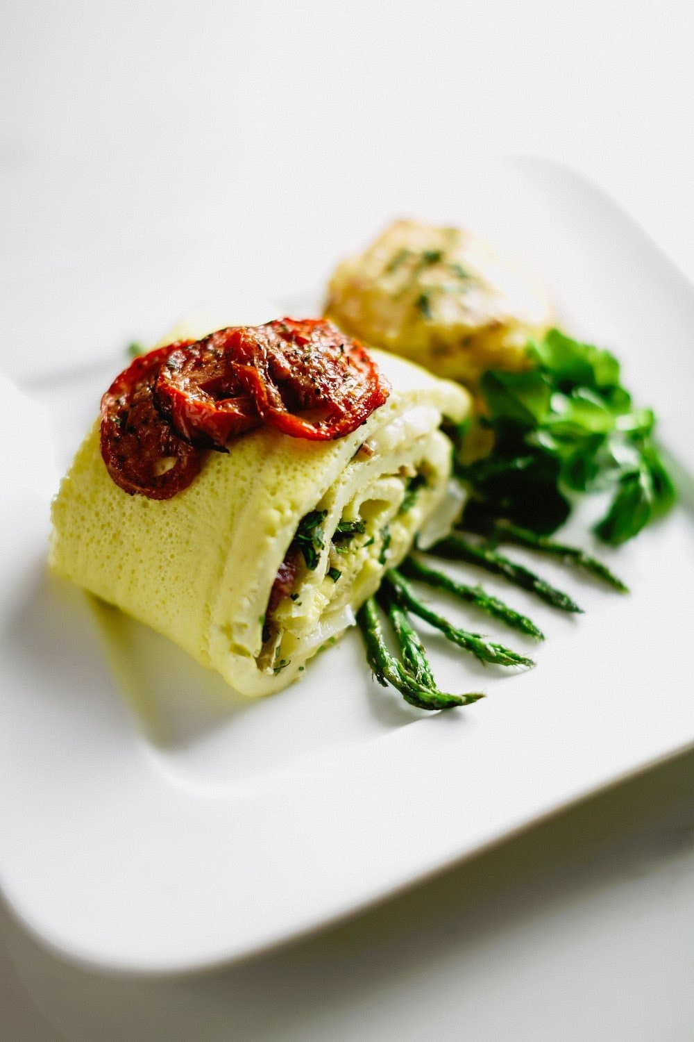 A rolled omelet filled with vegetables, topped with sun-dried tomatoes, served on a white plate with fresh greens and asparagus.