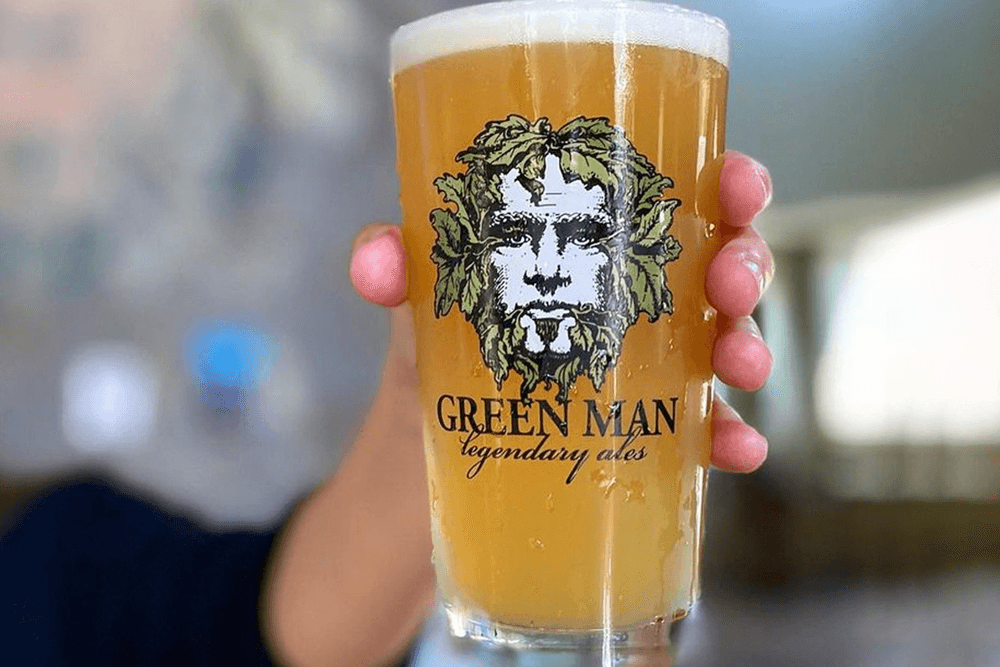 A hand holds a pint glass of beer featuring a "Green Man" logo.