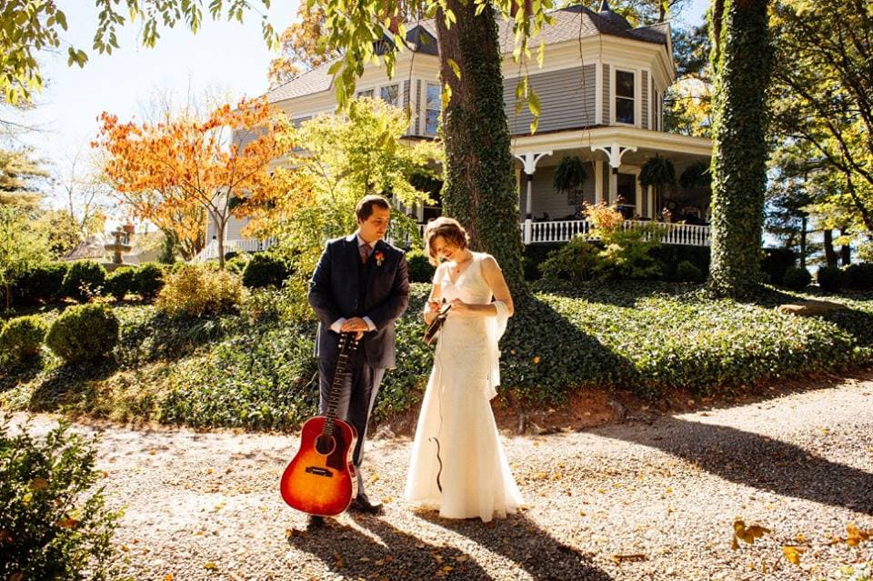 A couple stands together in a scenic garden, with the groom holding a guitar and the bride looking at her phone.