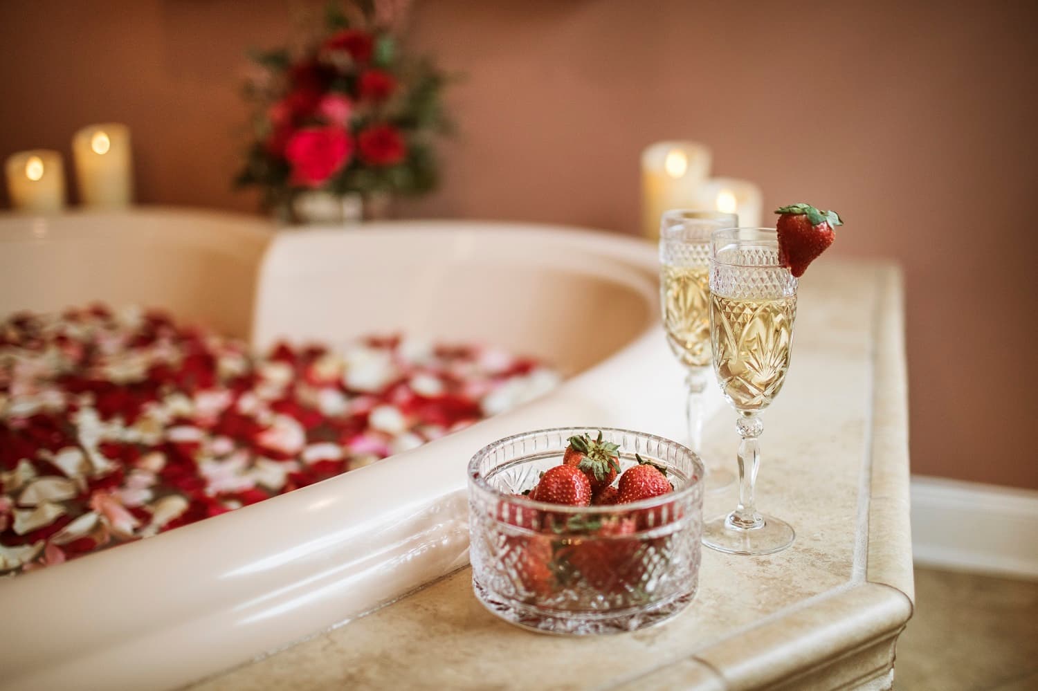 A luxurious spa setting featuring a bathtub filled with rose petals, two glasses of champagne, and a bowl of fresh strawberries.