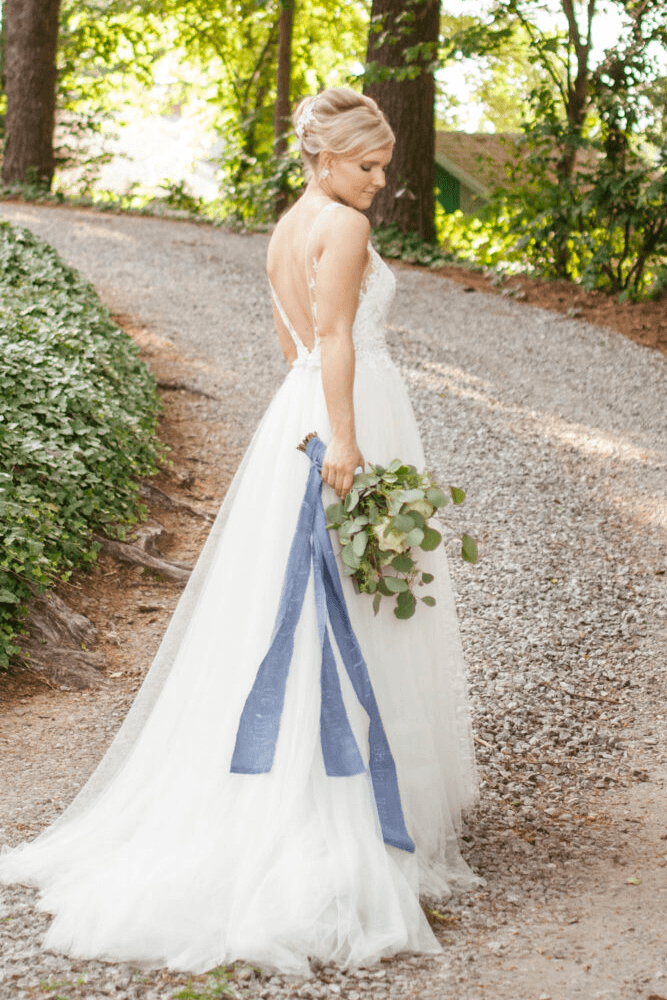 A bride walks along a gravel path, holding a bouquet and trailing a blue ribbon from her dress.
