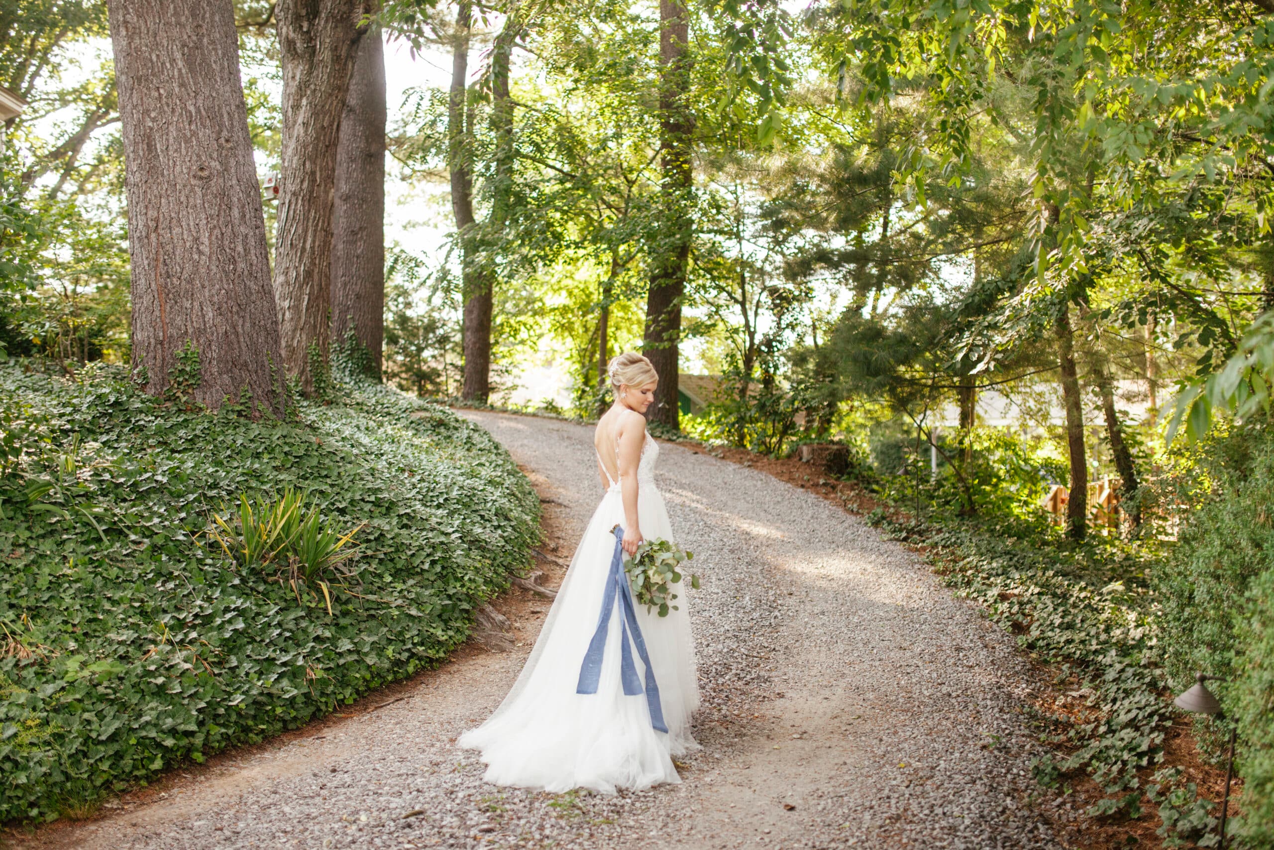 A bride in a white gown walks down a winding gravel path surrounded by lush greenery.