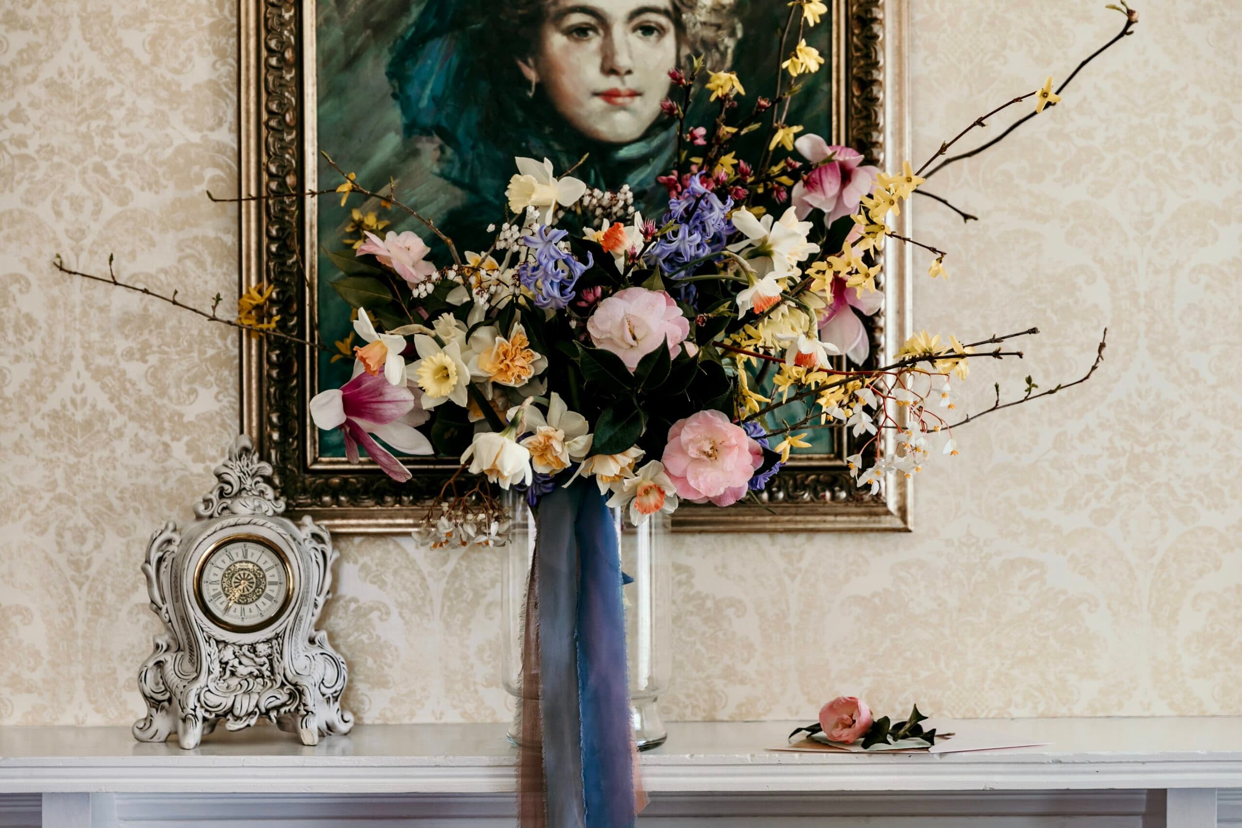 A vibrant floral arrangement in a vase sits on a mantel beneath a vintage portrait and clock.