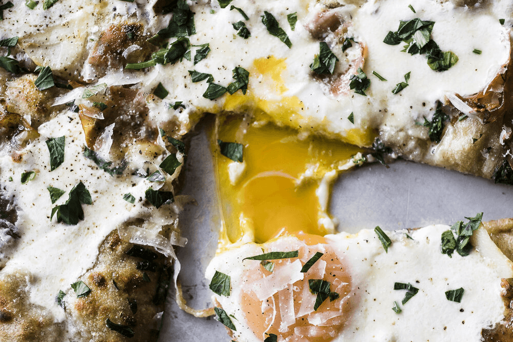 Close-up of a pizza topped with melted cheese, herbs, and a runny egg yolk.