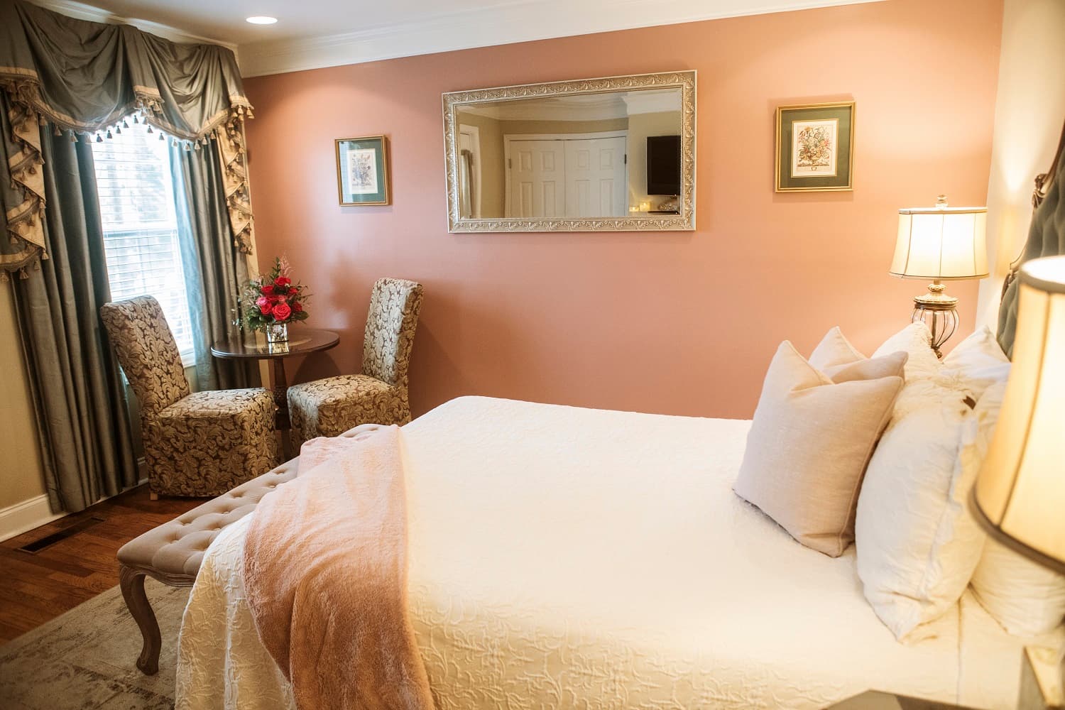 Cozy bedroom featuring a queen-sized bed, decorative pillows, and a small sitting area with two chairs and a table under a mirror.