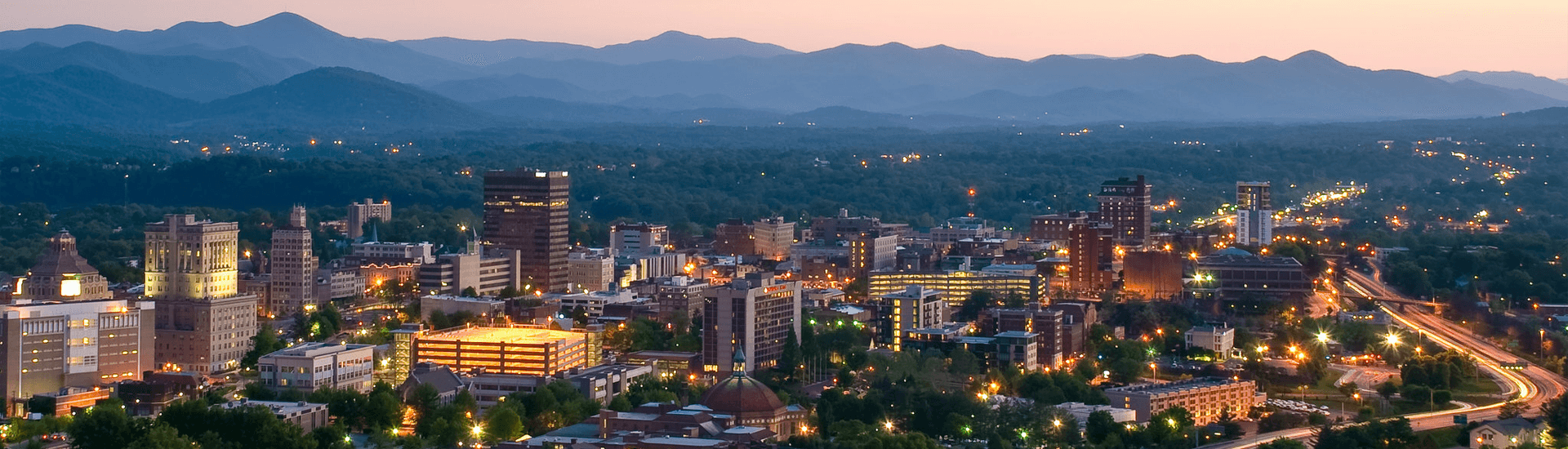 Panoramic view of a city skyline at dusk, surrounded by mountains.