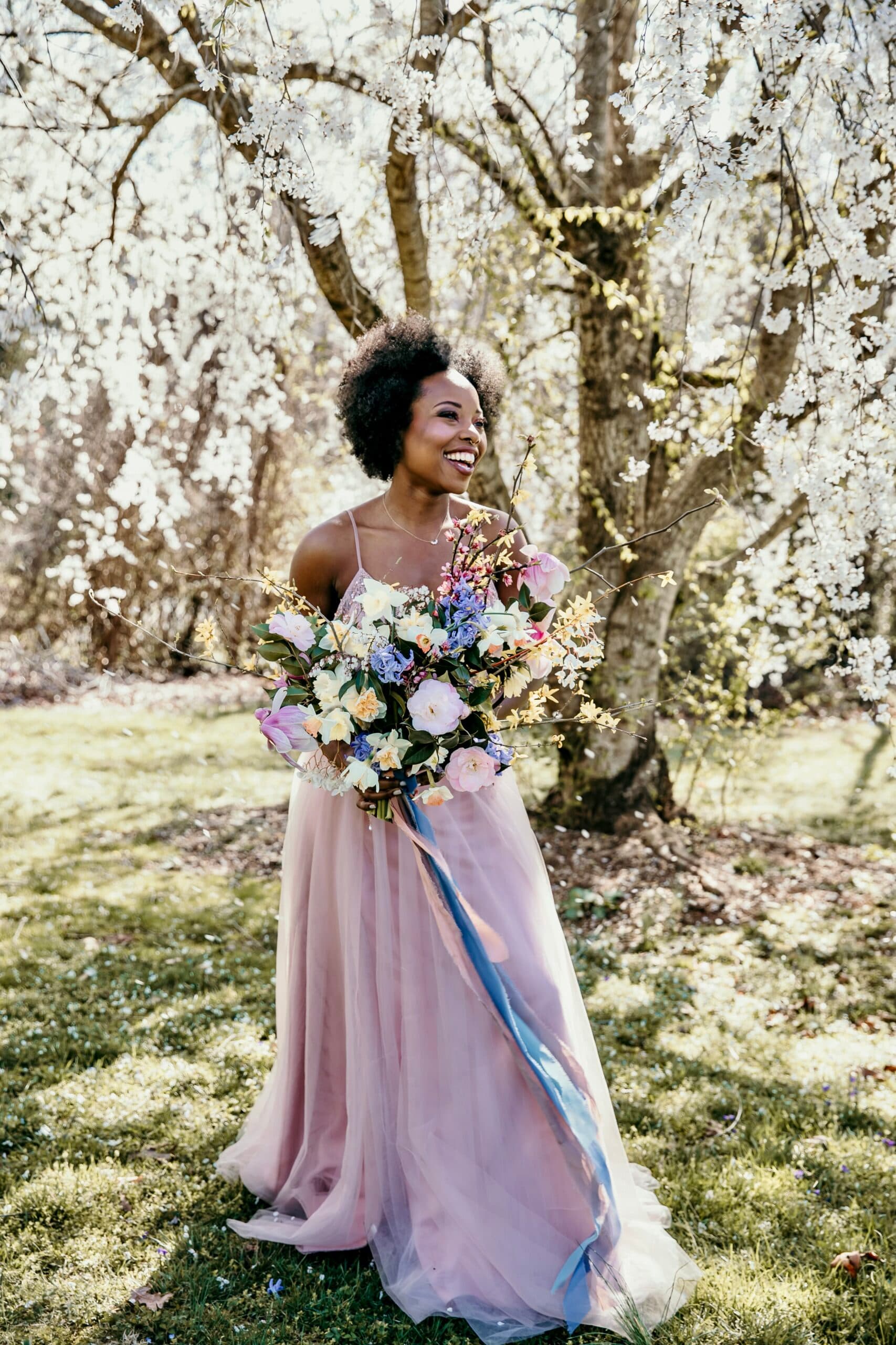 A woman in a flowing pink dress joyfully holds a vibrant bouquet under blooming trees.