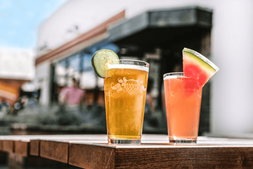 Two colorful drinks on a wooden table with outdoor seating in the background.