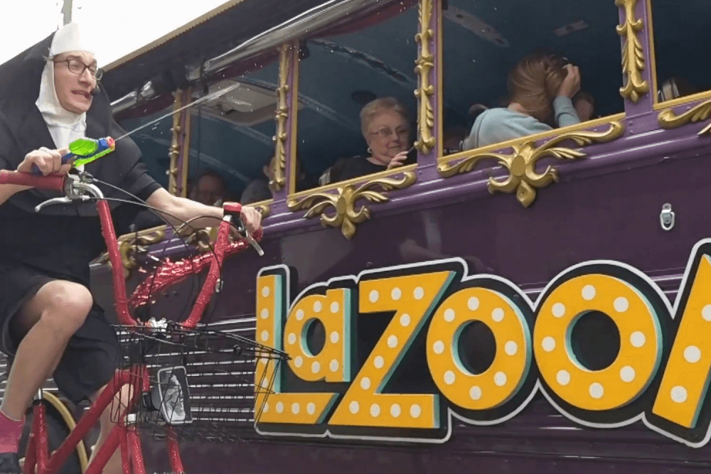 A person dressed as a nun rides a decorated bicycle while spraying water at amused passengers on a colorful bus.