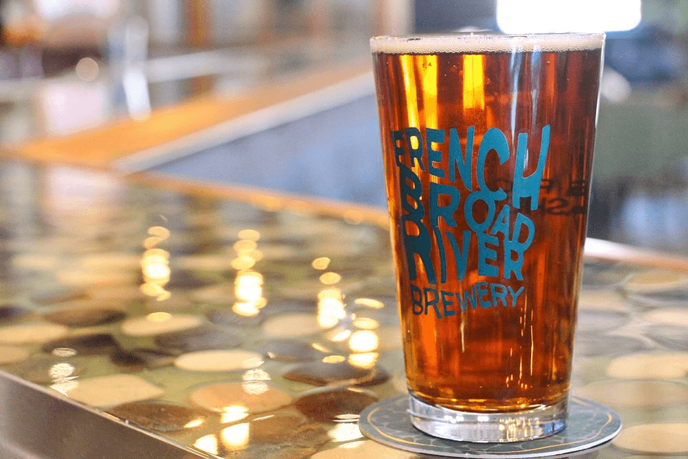 A pint of amber beer branded with "French Broad River Brewery" rests on a decorative table.