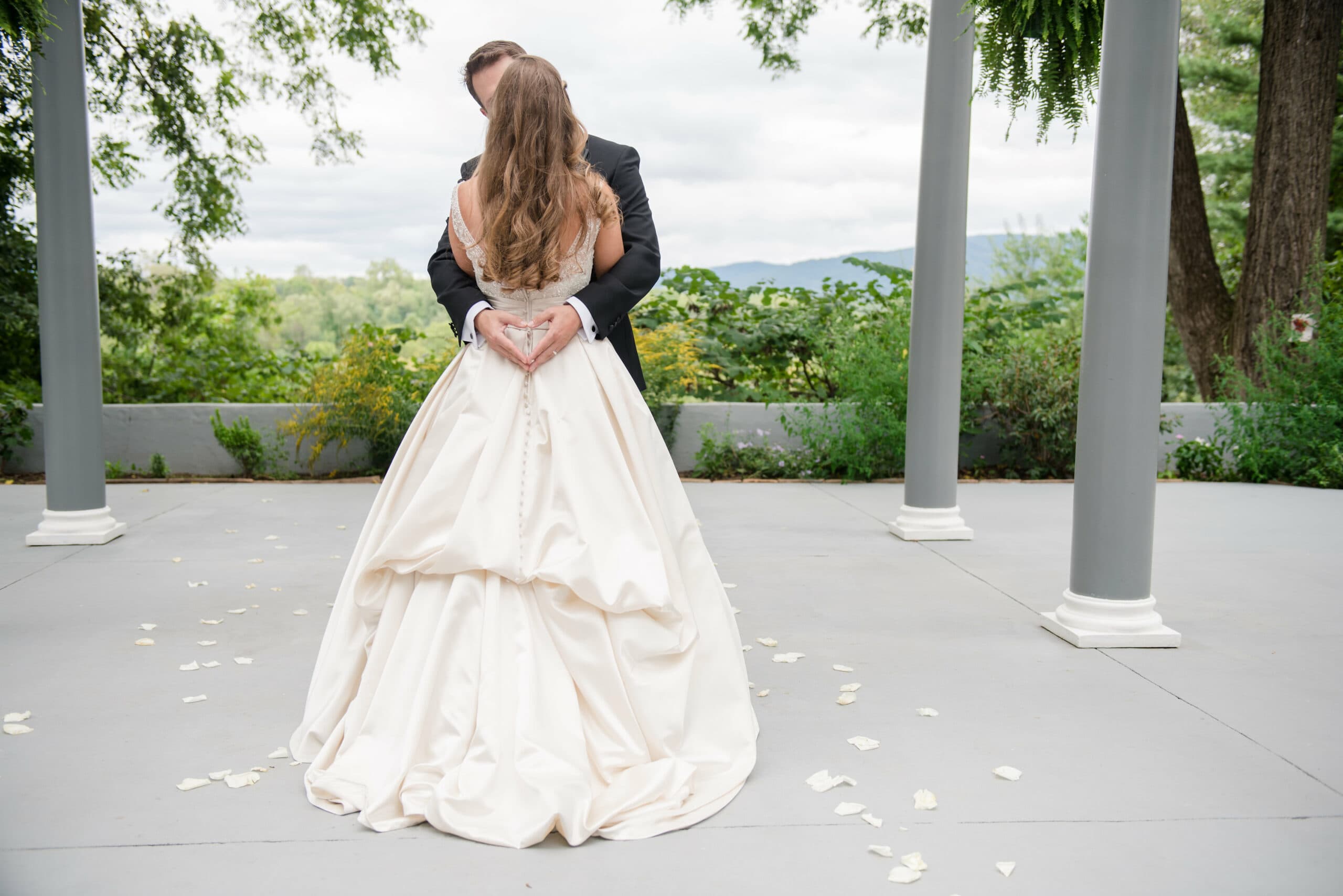 A bride and groom embrace outdoors, forming a heart shape with their hands, surrounded by nature.