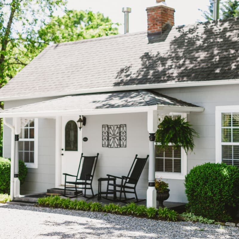 A charming gray house with a front porch featuring two black rocking chairs and lush green landscaping.
