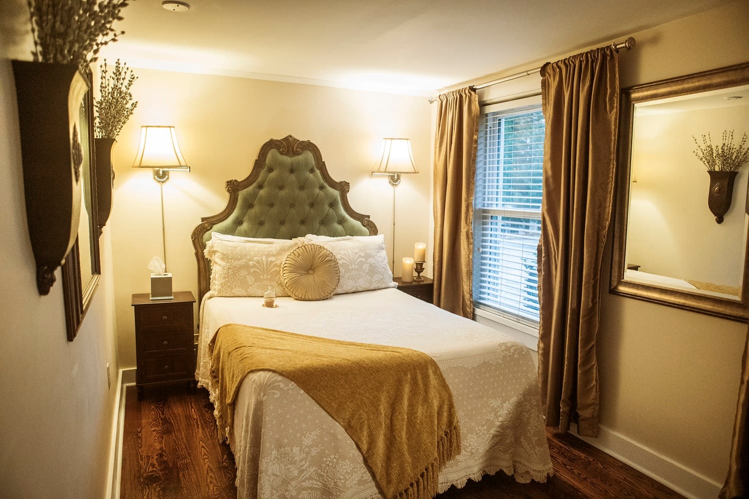 A cozy bedroom featuring a plush green headboard, decorative pillows, soft lighting, and warm-toned curtains.