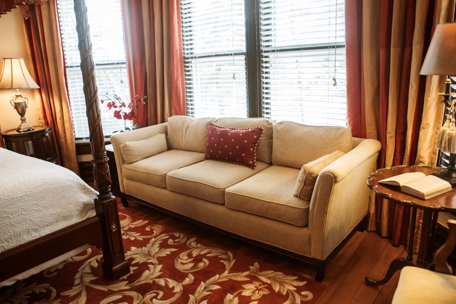 A cozy living area featuring a beige sofa with decorative pillows beside a wooden side table and a bed in a warmly decorated room.