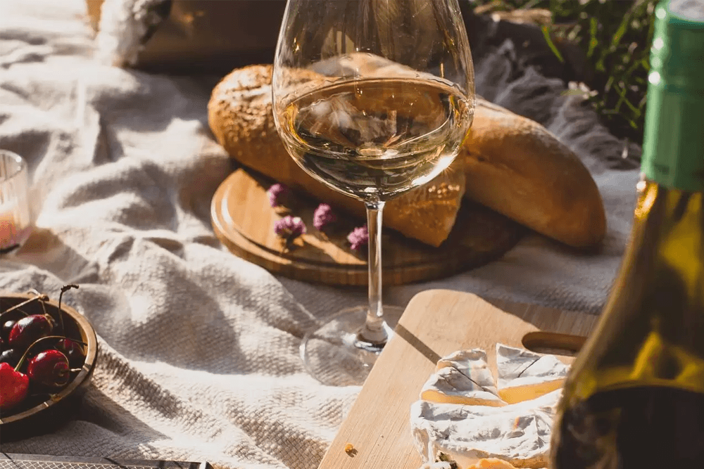 A glass of white wine sits beside bread, cheese, and cherries on a picnic blanket.