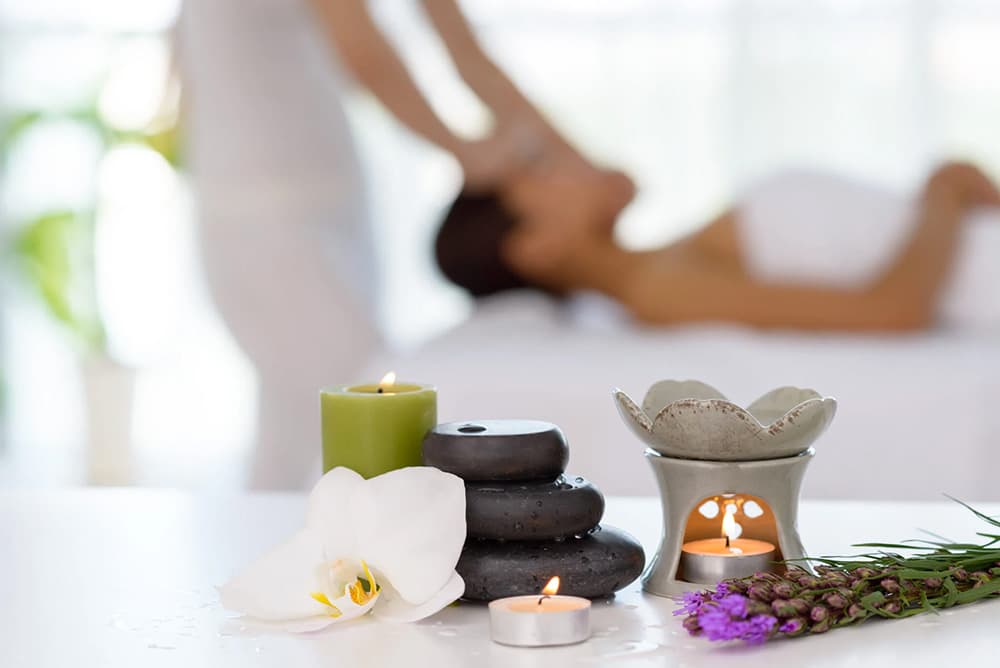 A serene spa setting featuring massage stones, candles, and an orchid, with a person receiving a massage in the background.