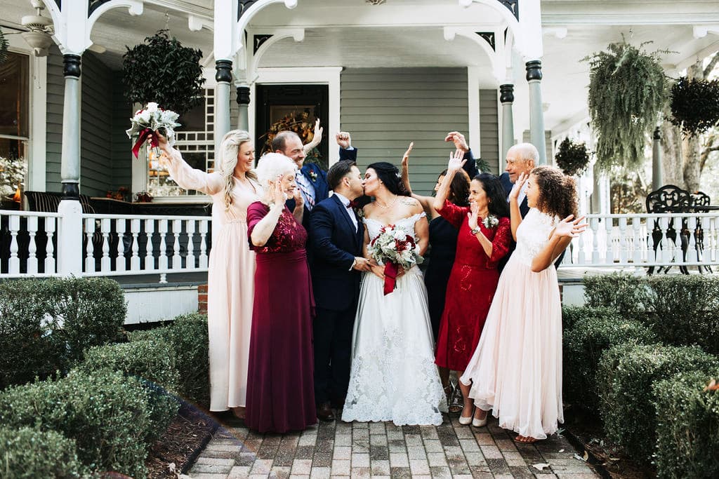 A joyful bridal party celebrates the couple's kiss outside a charming house.